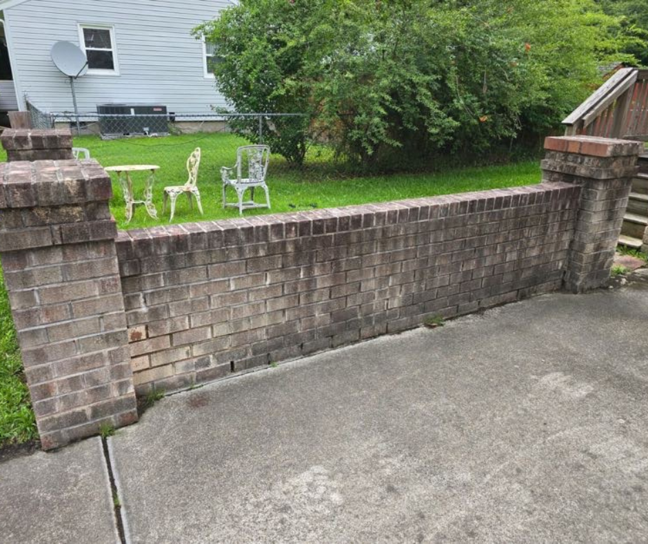 Brick retaining wall and concrete driveway showing dirt, algae, and weather staining before professional pressure washing, with a residential backyard visible in the background.