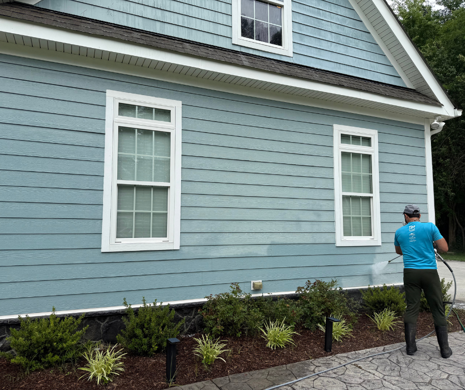 Technician soft washing the exterior siding of a blue home to remove dirt, algae, and mildew during a professional house washing service.