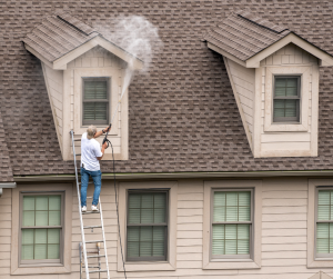 Technician pressure washing roof shingles and dormer siding from a ladder