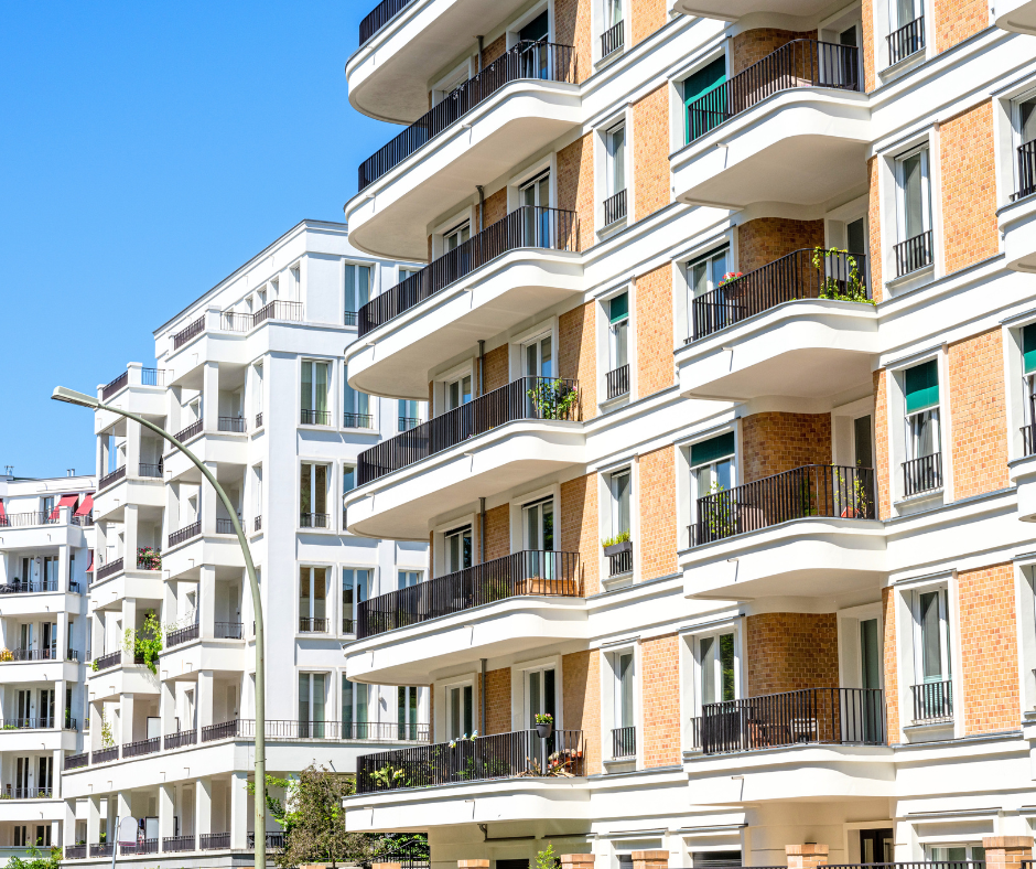 Modern multi-story apartment building with brick and white exterior walls, curved balconies, black railings, and landscaped terraces under a clear blue sky.